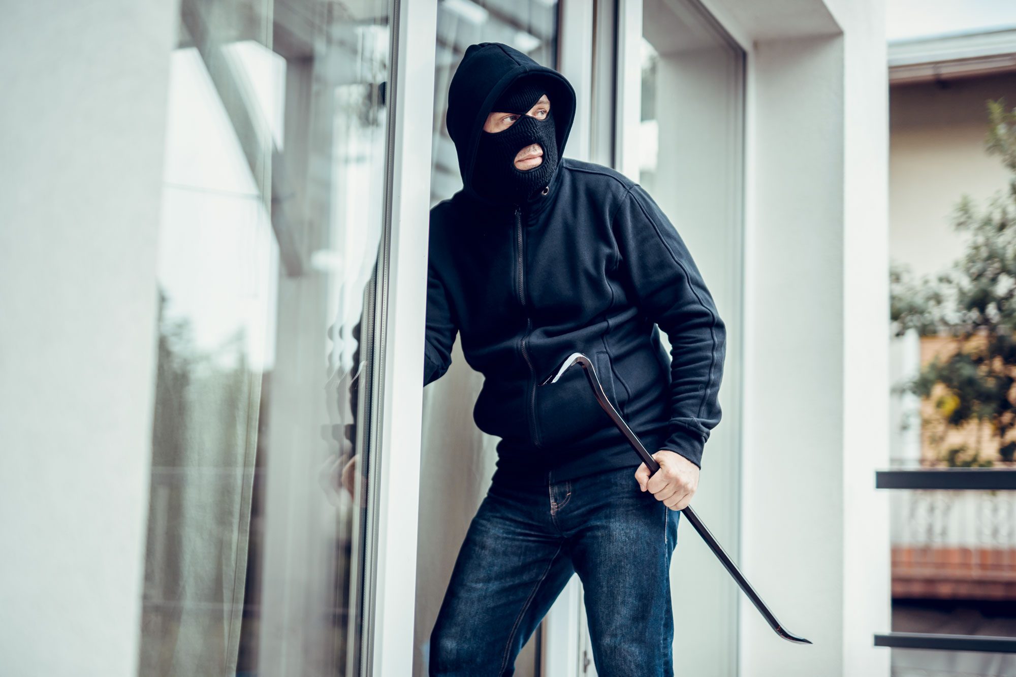 Masked figure holds crowbar by glass door, appearing to attempt entry, with a blurred outdoor background.