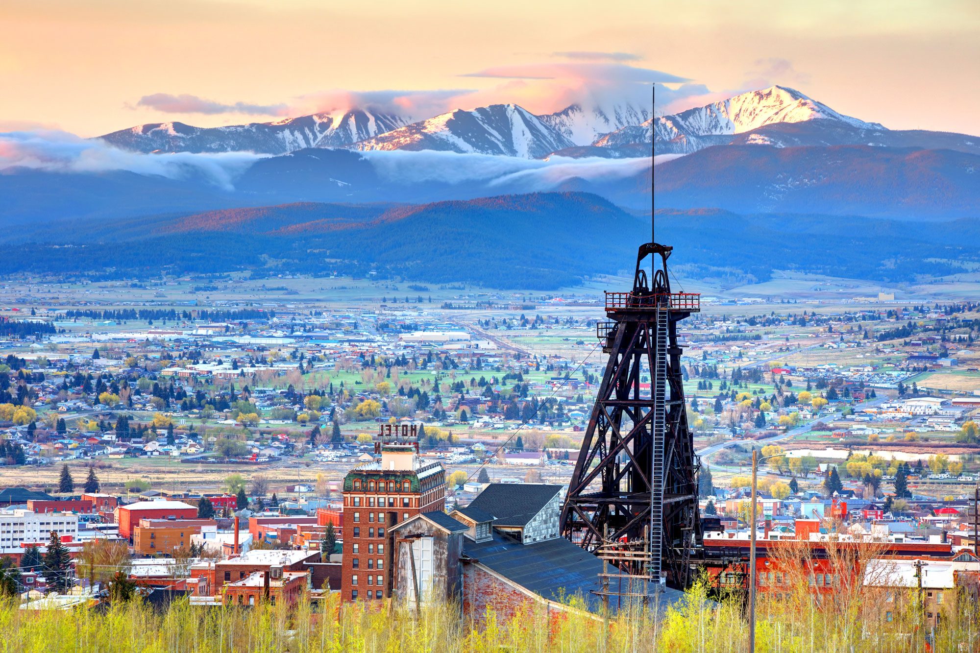 Industrial structure stands over cityscape, framed by snow-capped mountains and sunrise-lit clouds in the distance. "MOTEL" sign visible on a nearby building.