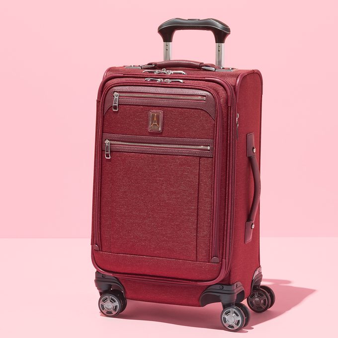 Red suitcase stands upright on four wheels in a light pink setting.