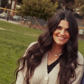 A person smiles, standing outside on a grassy field with trees in the background.
