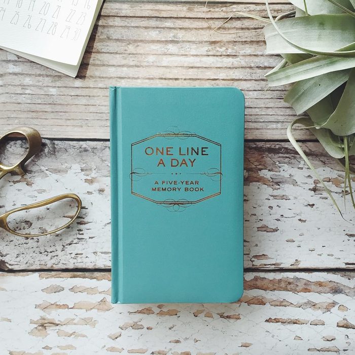 A teal memory book lies on a rustic wooden surface next to a plant, calendar pages, and brass scissors.