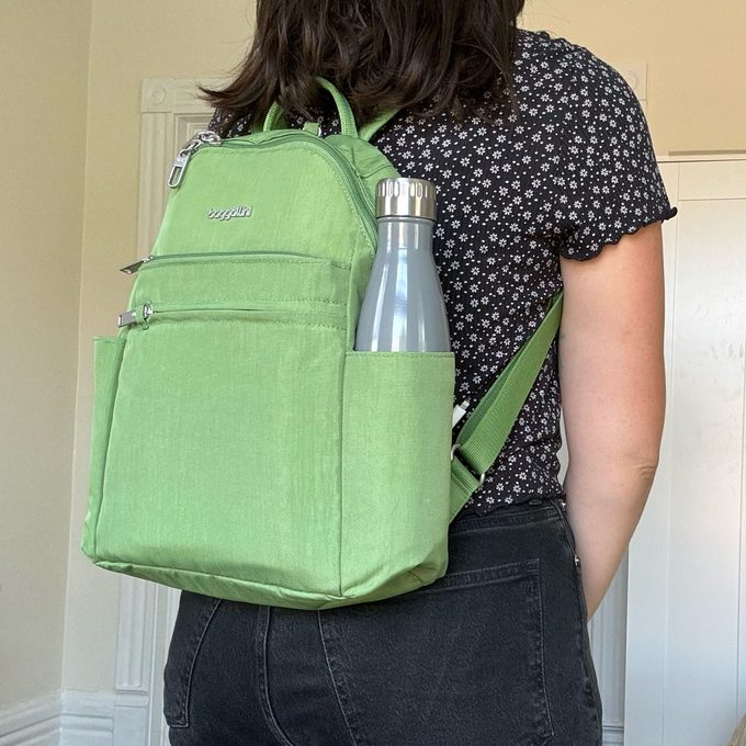 Backpack holds water bottle, worn by person standing indoors, against a light-colored wall.