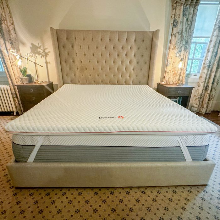 Bed displays a Dormeo mattress topper in a cozy bedroom with floral curtains and bedside tables holding lamps.