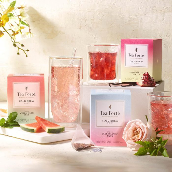 Tea sachets sit with cold brew glasses and fruit slices, surrounded by tea boxes. Bright, floral setting with pomegranate and rose accents.