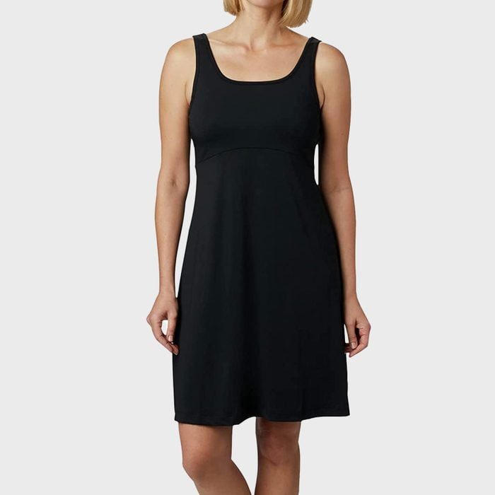 A woman stands wearing a sleeveless black dress against a plain gray background.