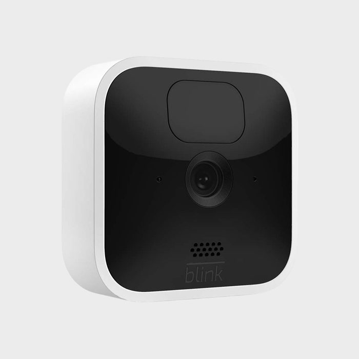 A compact, square black-and-white security camera with a single lens sits against a plain gray background. 