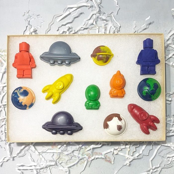 Colorful, space-themed crayons displayed in a foam-lined box with white shredded paper surrounding them.