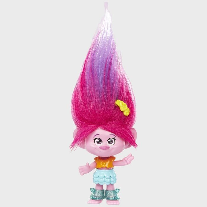 A troll doll stands, featuring vibrant pink hair, a yellow hairpin, and wearing colorful clothing in a simple studio setting.