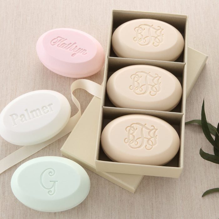 Soap bars display engravings, resting on a wooden surface beside a box and ribbon. Text includes 