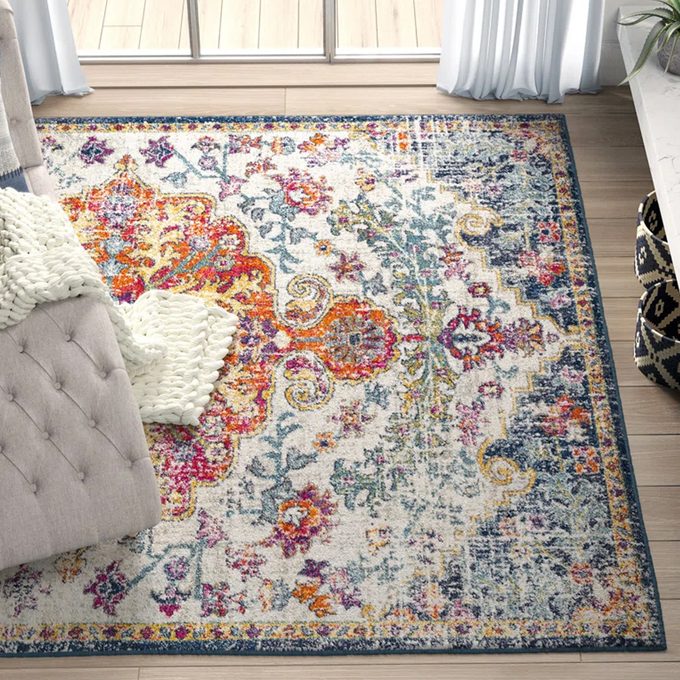 A colorful, patterned rug lies on a wooden floor beside a tufted sofa and a knitted white blanket near large windows and curtains.