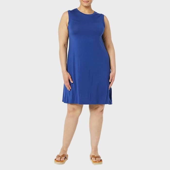 A person stands wearing a sleeveless blue dress, brown sandals, against a plain light gray background.