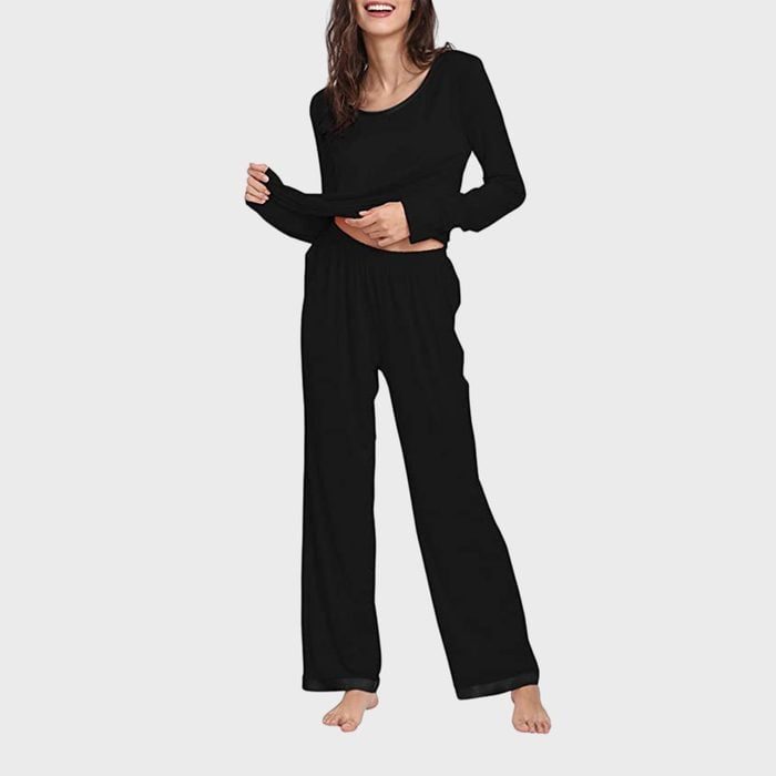 Woman wearing black loungewear stands barefoot, slightly lifting her shirt, against a plain light gray background.