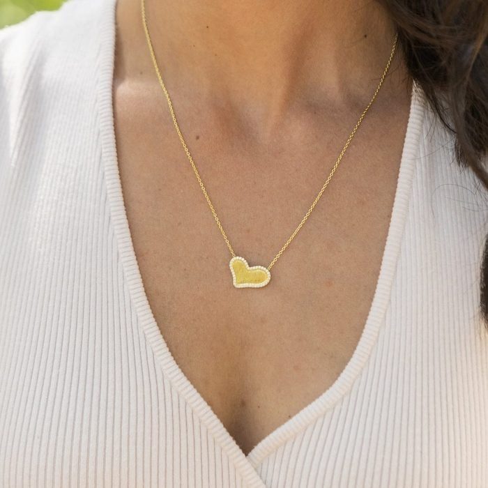 Gold heart pendant rests on a gold chain, worn with a ribbed, light-colored V-neck top, with blurred greenery in the background.