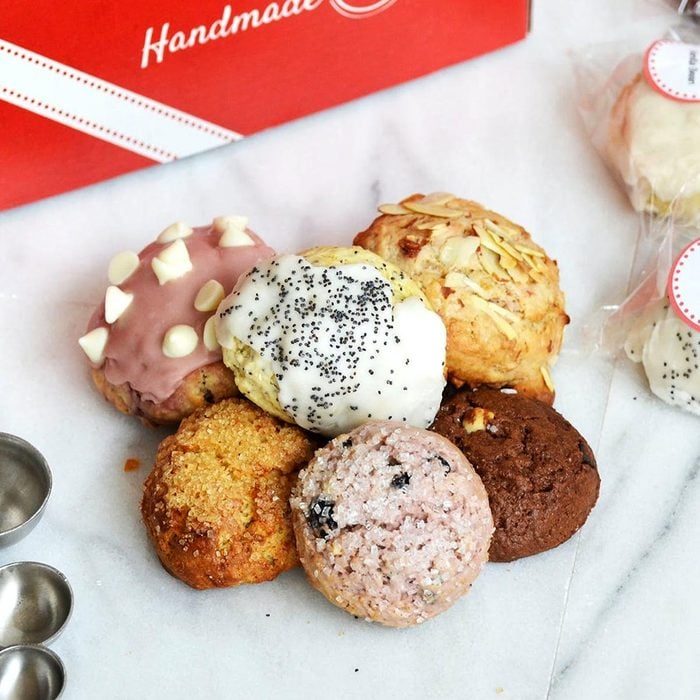 Cookies rest arranged in various flavors and toppings; set on a marble surface, next to a red box labeled 