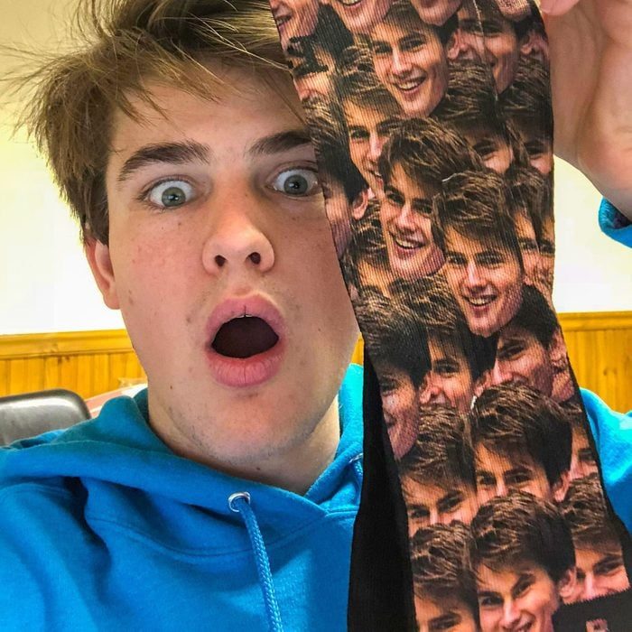 Person holds a sock featuring a pattern of repeated faces, looking surprised. Background is a room with wooden paneling.