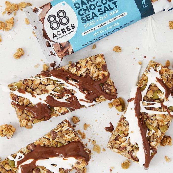 Granola bars drizzled with chocolate and white frosting rest on a white surface, accompanied by scattered granola crumbs and product packaging.
