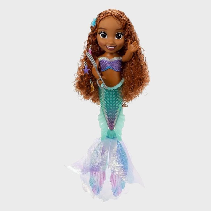 Mermaid doll waves, holding seashell accessory; wearing shimmering purple and teal outfit, surrounded by simple white background.