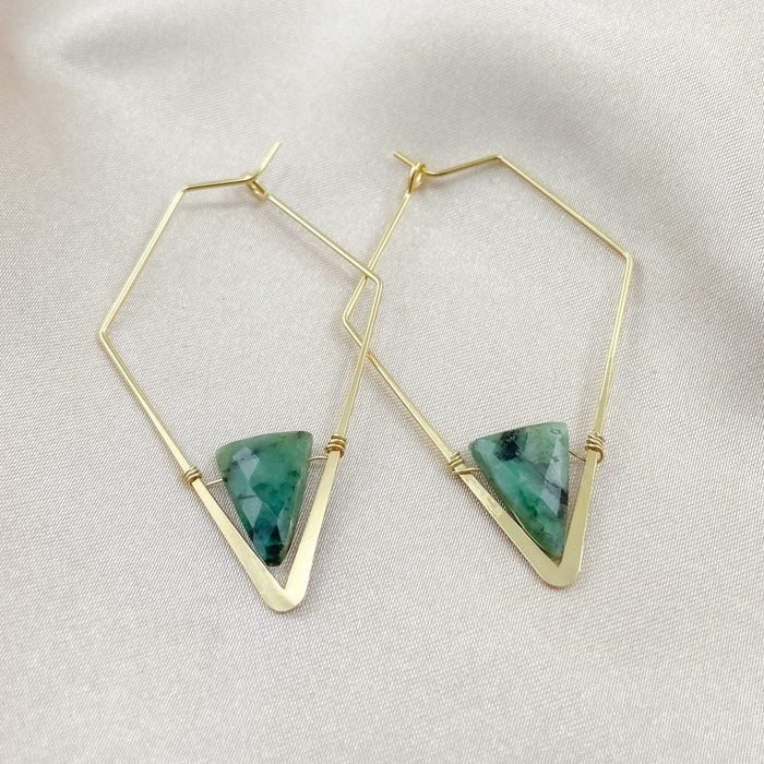 Gold hexagonal earrings feature green triangular gemstones, resting on smooth, light fabric.