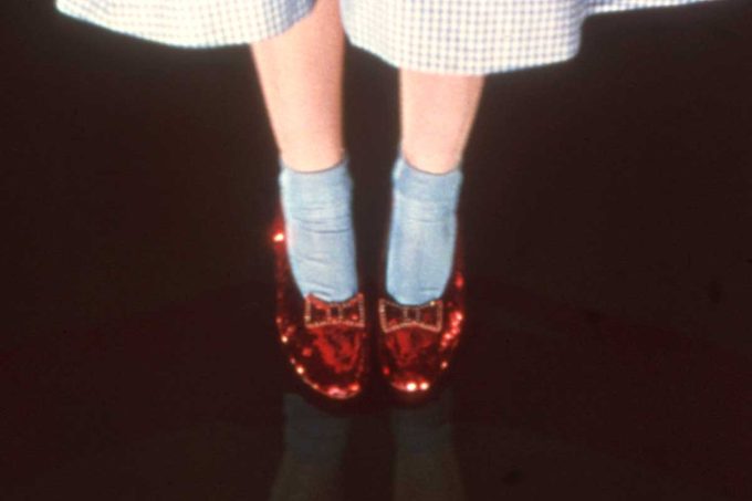 Red shoes sparkle, worn by feet in blue socks, under a gingham dress, standing on a dark reflective surface.