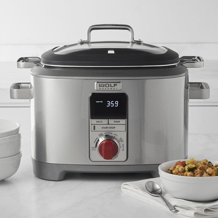 Stainless steel slow cooker displaying a timer, sits on a kitchen countertop beside stacked bowls and a bowl of stew with a spoon.