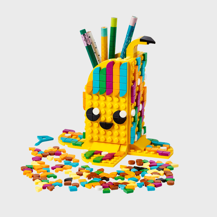 A colorful LEGO pencil holder with a smiling face holds pencils, surrounded by scattered multicolored LEGO pieces on a plain background.