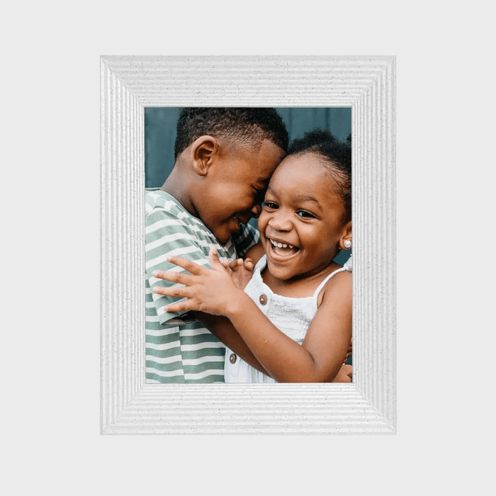 Two children embrace joyfully, smiling widely. One wears a striped shirt, the other a white dress, inside a textured, white picture frame.