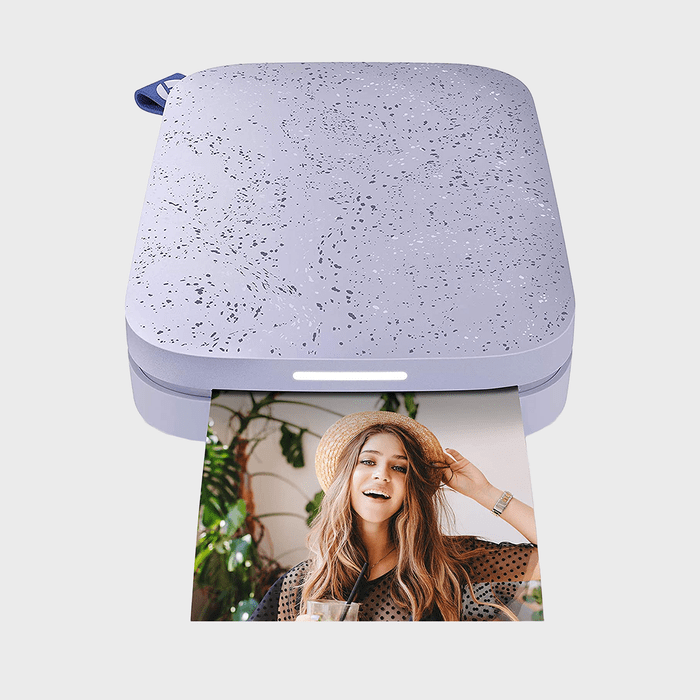 Printer ejects a photo of a smiling woman wearing a hat. Background features plants. Device appears sleek with a speckled surface.
