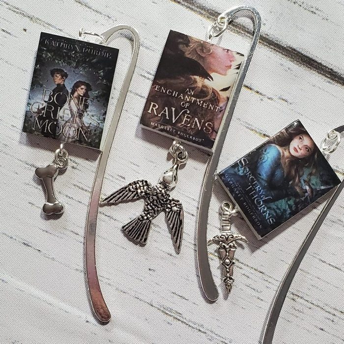 Bookmarks display miniature book covers with charms; a bone, bird, and sword. They rest on a light, textured surface.