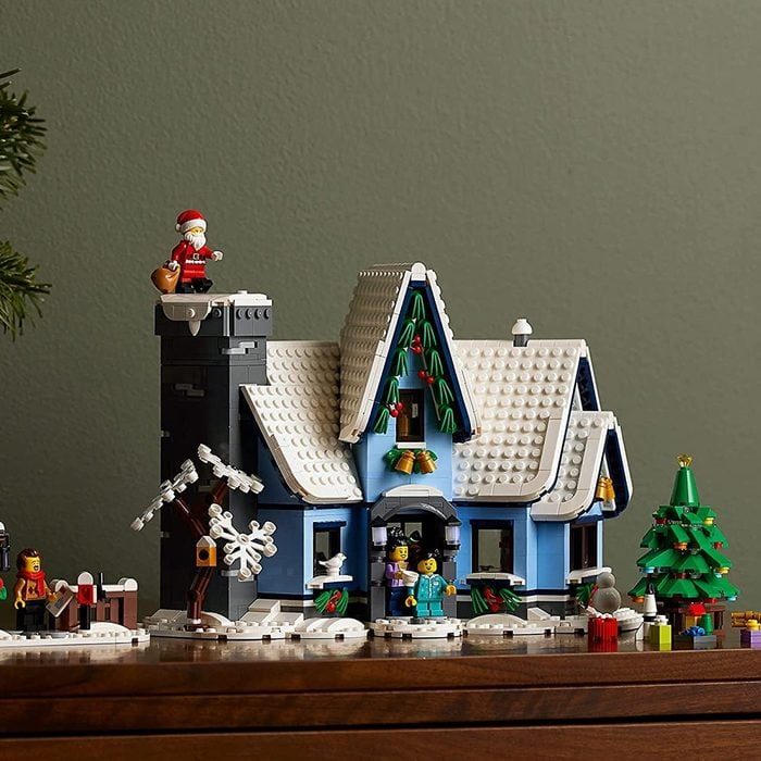 Lego house sits festively decorated, featuring Santa on chimney, surrounded by trees and gifts on a wooden table against a green wall.
