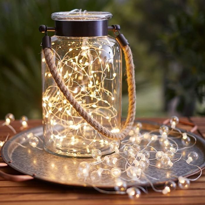 A glass jar contains glowing fairy lights, sitting on a metal tray, surrounded by a wooden table outdoors.