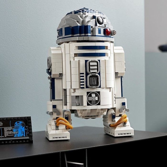 LEGO robot model stands on a dark surface, detailed with blue and white bricks, in a light-colored room. Nearby is a display plaque.