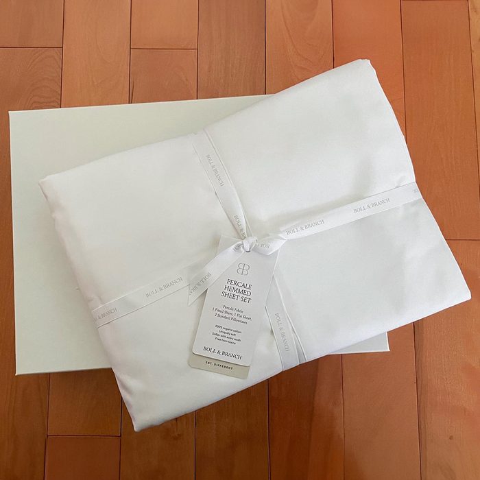 White sheet set lies folded, tied with branded ribbon on wood floor, labeled 