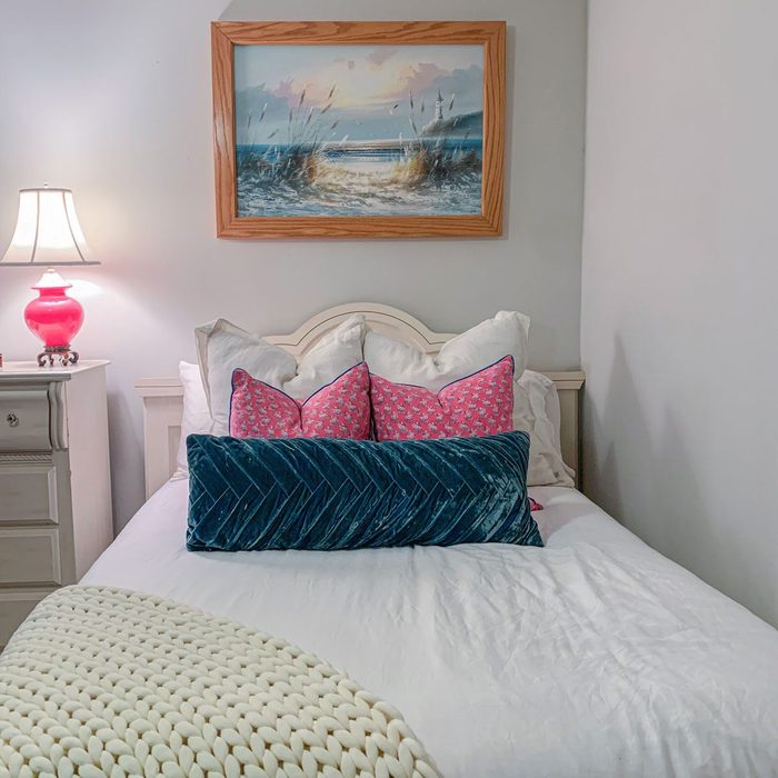 Bed is neatly arranged with colorful pillows, placed under a seascape painting. A nightstand holding a lamp completes the cozy bedroom setting.