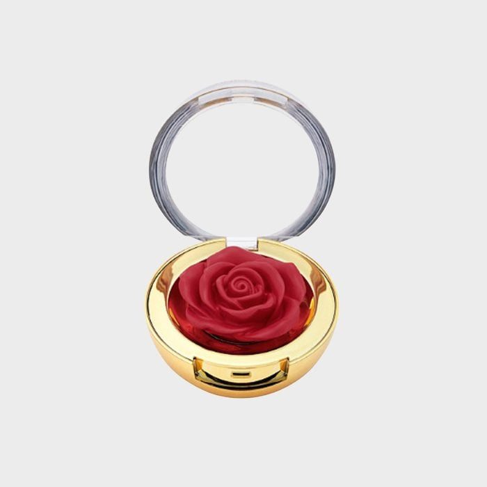 A rose-shaped cosmetic compact is open, displaying a vivid red rose blush nestled inside a golden case against a plain gray background.
