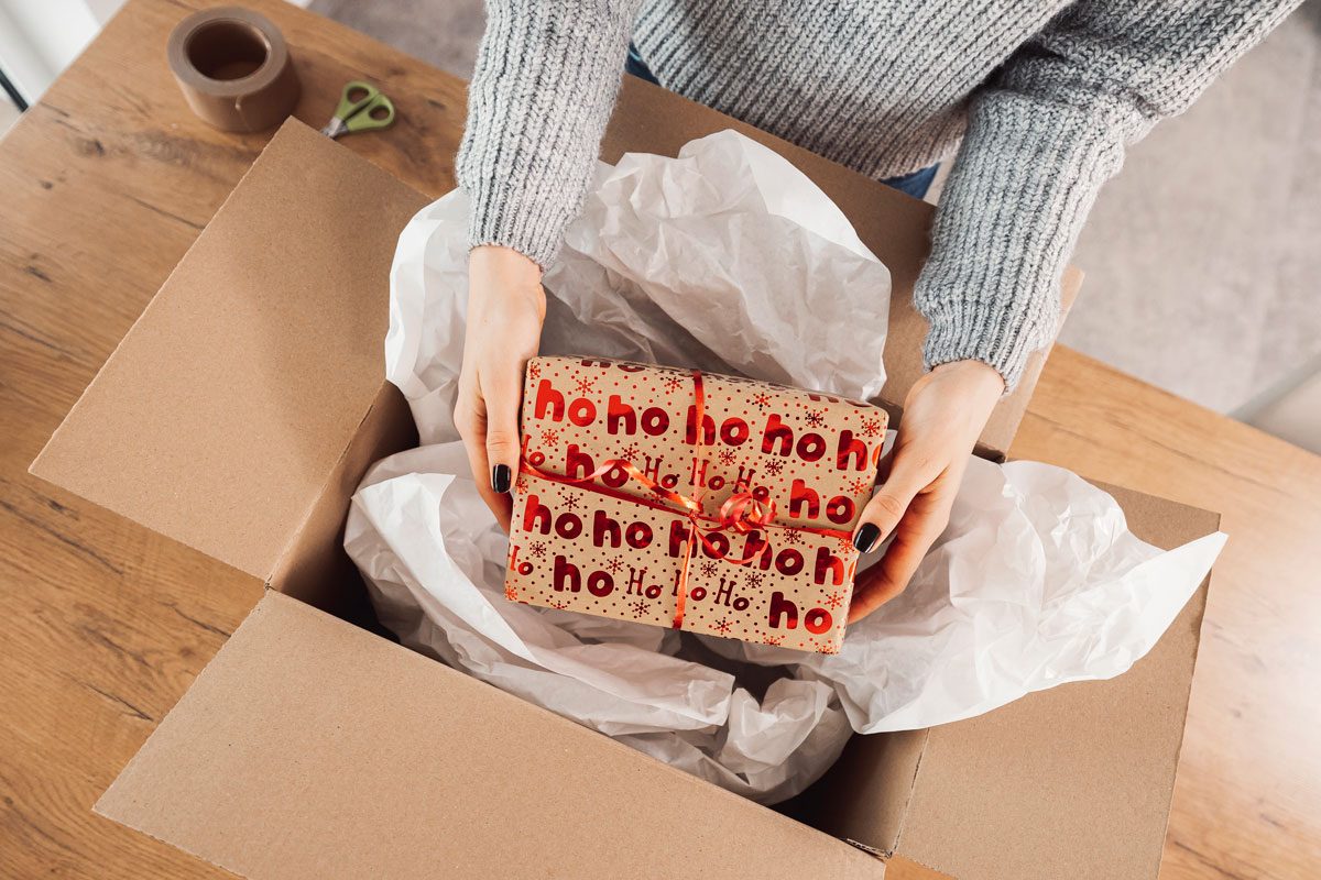 Hands place gift wrapped in "ho ho ho" paper into a cardboard box filled with tissue paper on a wooden table.