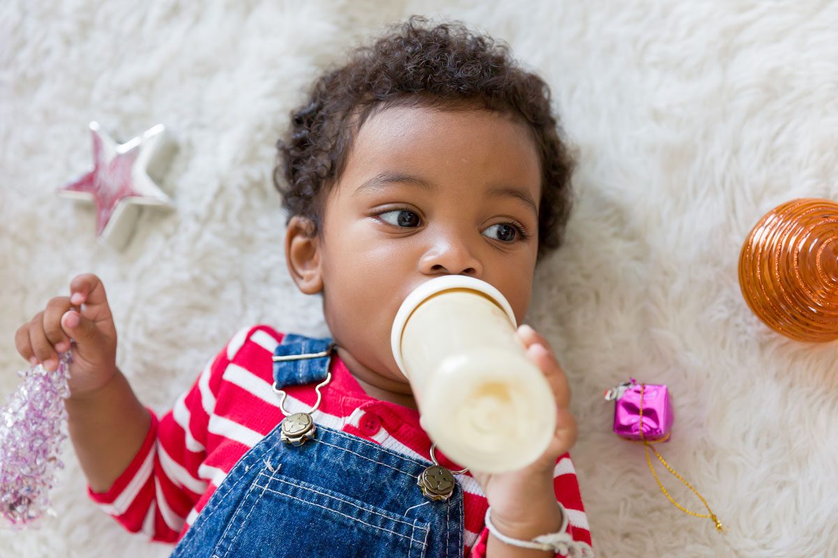 African American Little Boy Lying On Floor And Eating Milk From Nipples Bottle