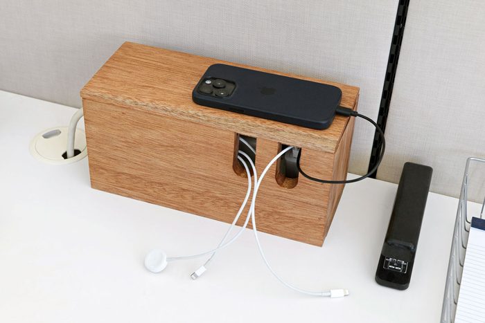 a wooden cable management box on a white desk. Various cords and cables are plugged into the box, including charging cables for a smartphone and headphones; The box has several openings for cables to pass through, helping to organize and conceal the cords