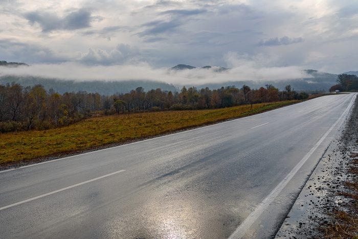 Bright picturesque view from the asphalt road, wet after the rain, mountains, fog, golden trees and grass against a blue sky with clouds