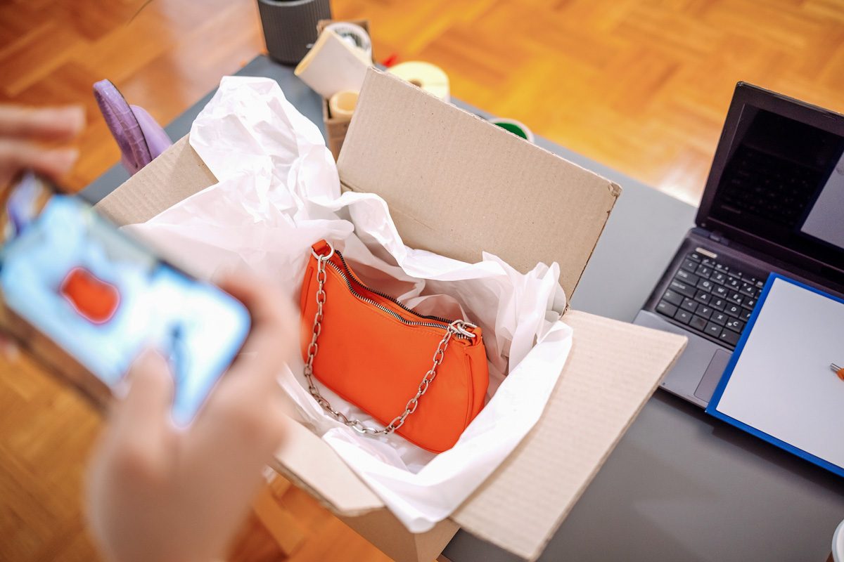 woman taking a picture of a bag in opened cardboard box next to laptop